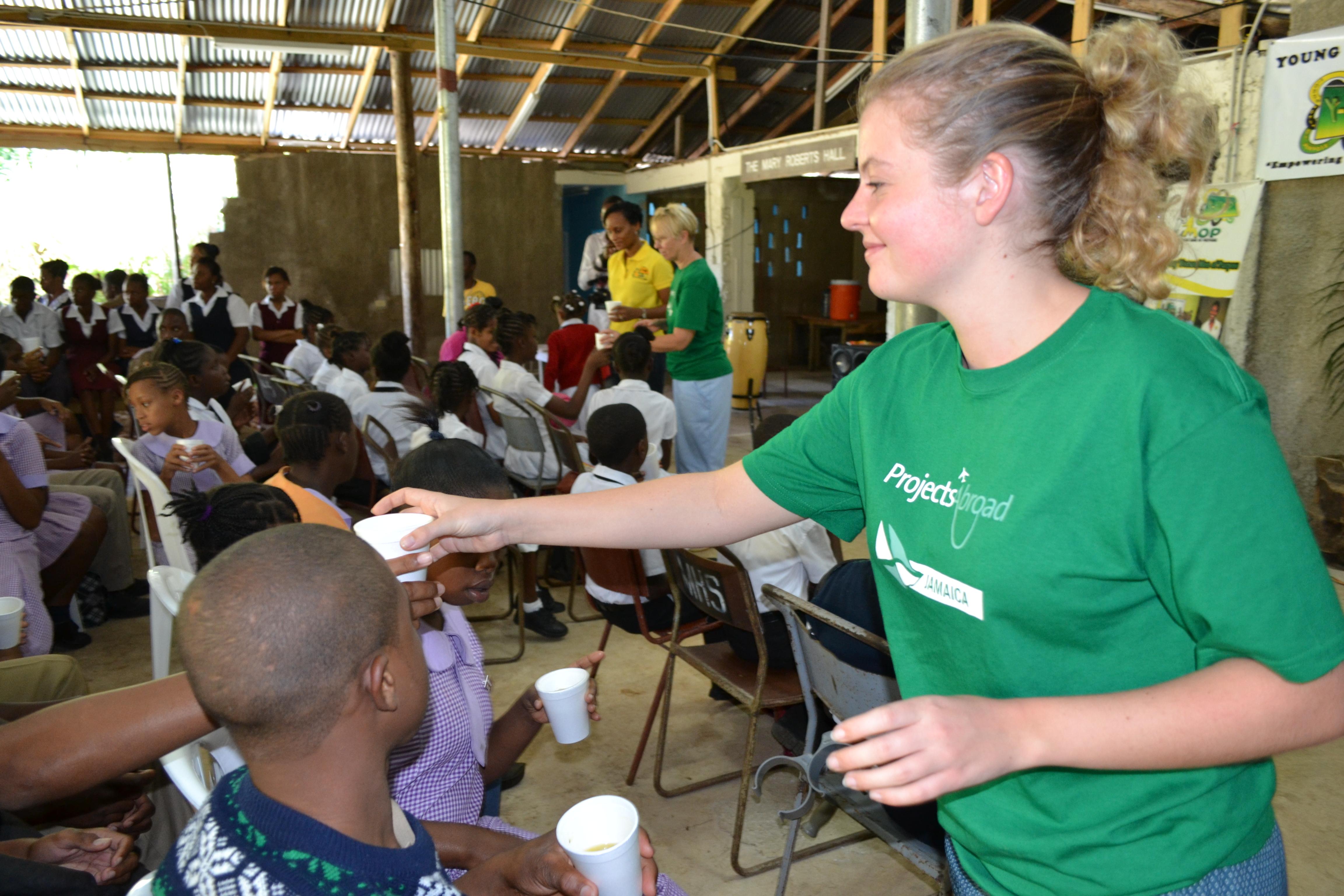 Projects Abroad Childcare volunteer in Jamaica is handing out hot drinks at a local school during a HIV/Aids awareness presentation. Psychology work experience in Jamaica shows Projects Abroad Childcare volunteer handing out hot drinks to school children during a HIV/Aids awareness presentation.